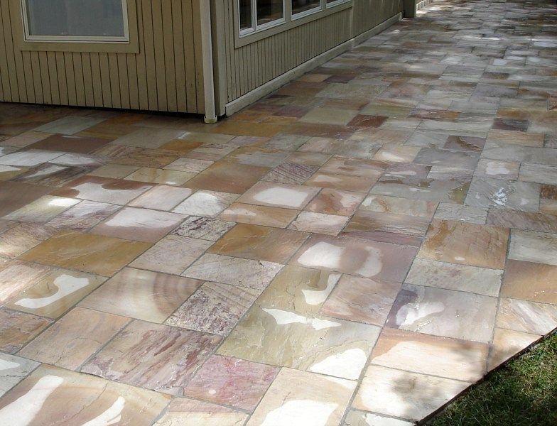 A stone patio with rectangular pavers in varied shades of tan, brown, and pink, adjacent to a beige house exterior.
