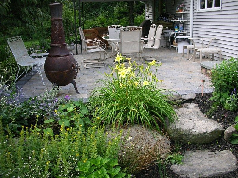 A patio with outdoor dining furniture and a tall, metal chiminea, surrounded by a garden with blooming yellow daylilies.