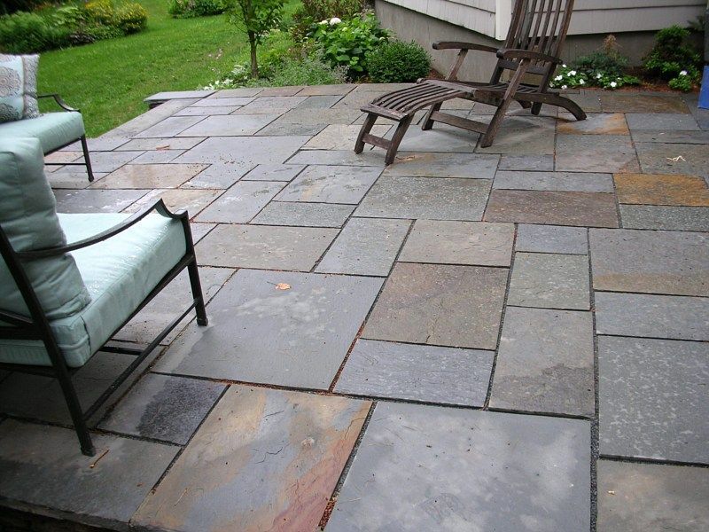 A stone patio with outdoor chairs and an ottoman on a sunny day next to a grassy lawn.