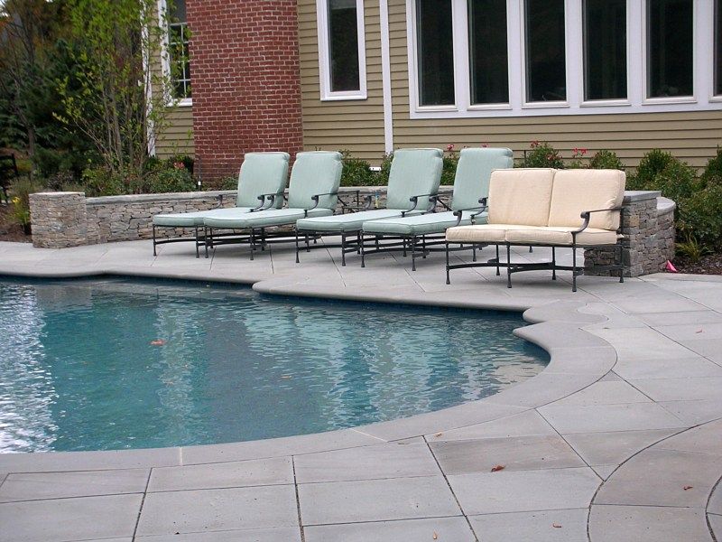 A swimming pool patio featuring four light green lounge chairs and a beige loveseat arranged against a stone retaining wall.