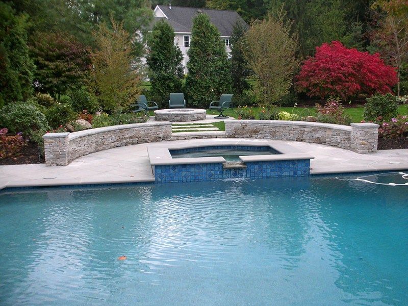 A blue swimming pool with a stone hot tub, a curved stone retaining wall, and a fire pit area in a backyard setting.