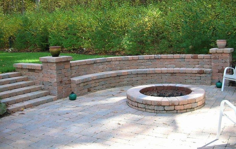 A stone patio featuring a circular fire pit and a curved built-in bench wall with stone stairs on the left.