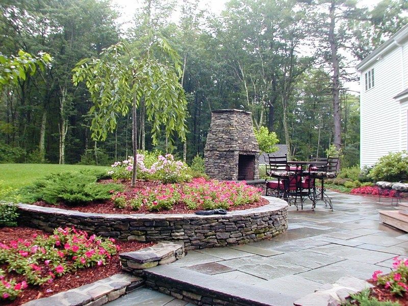 A stone fireplace and patio area with a tiered garden, pink flowers, and a small tree in a wooded backyard.