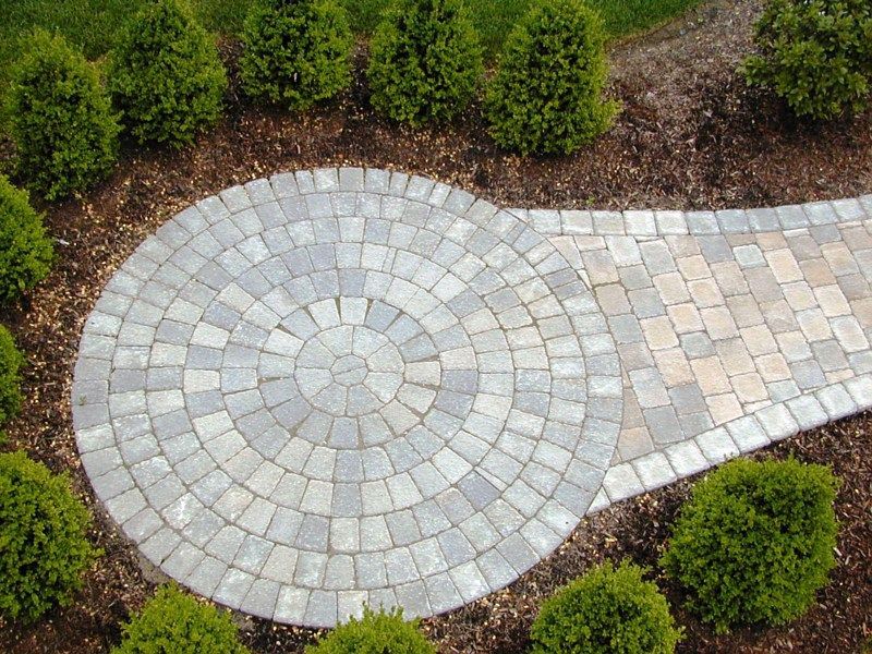 A circular paver patio with a radial pattern joins a rectangular walkway, surrounded by a mulch bed and small green bushes.