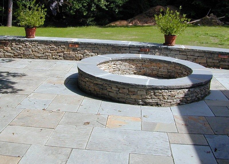 A circular stone fire pit sits on a stone patio in front of a low retaining wall with potted plants.