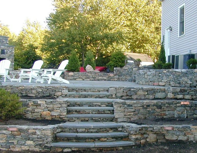 A tiered stone patio with steps leading to a seating area with white chairs, set against a house and lush green trees.