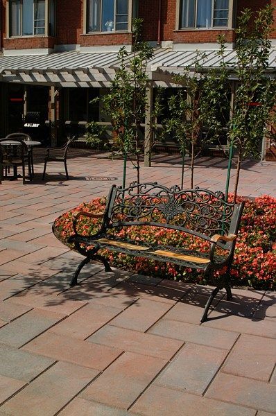 A decorative metal bench sits on a paved patio in front of a brick building with a pergola, surrounded by red flowers.
