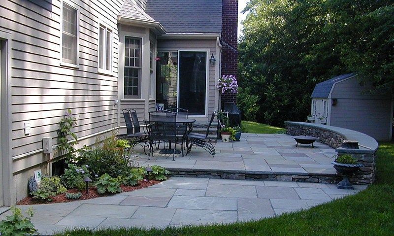 A gray stone patio with a dining set and a curved retaining wall attached to the back of a light-colored shingled house.