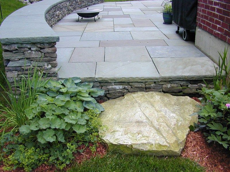 A flagstone patio with a curved stone sitting wall, a fire pit, and a large natural stone step in a landscaped yard.