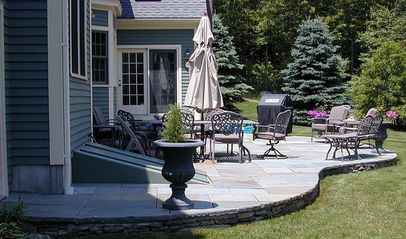 A flagstone patio with outdoor furniture, a table, and an umbrella next to a blue house with a cellar door.