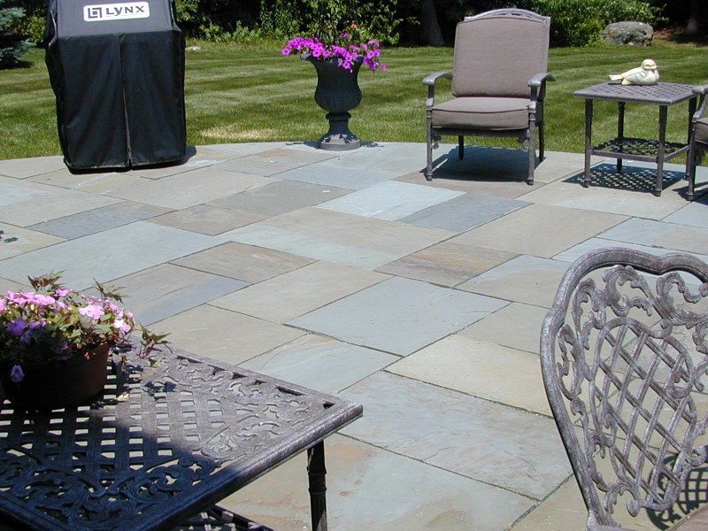 A patio with stone pavers, outdoor furniture, a grill, and planters with pink flowers on a sunny day.