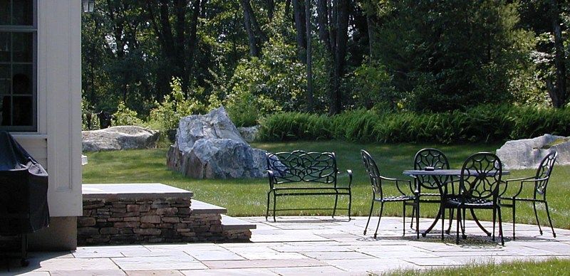 A flagstone patio featuring black wrought iron furniture, set in a backyard with a stone wall and large landscape boulders.