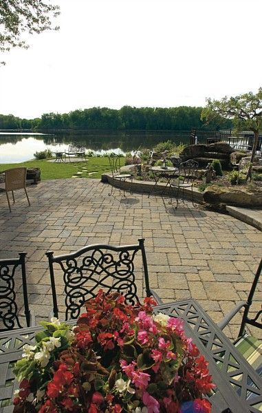A patio with dining chairs and a flower centerpiece overlooks a calm lake surrounded by trees on a bright, sunny day.