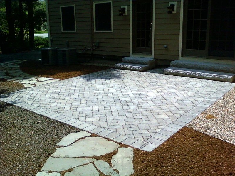 A new rectangular stone paver patio sits against the back of a house, featuring a herringbone pattern and border.