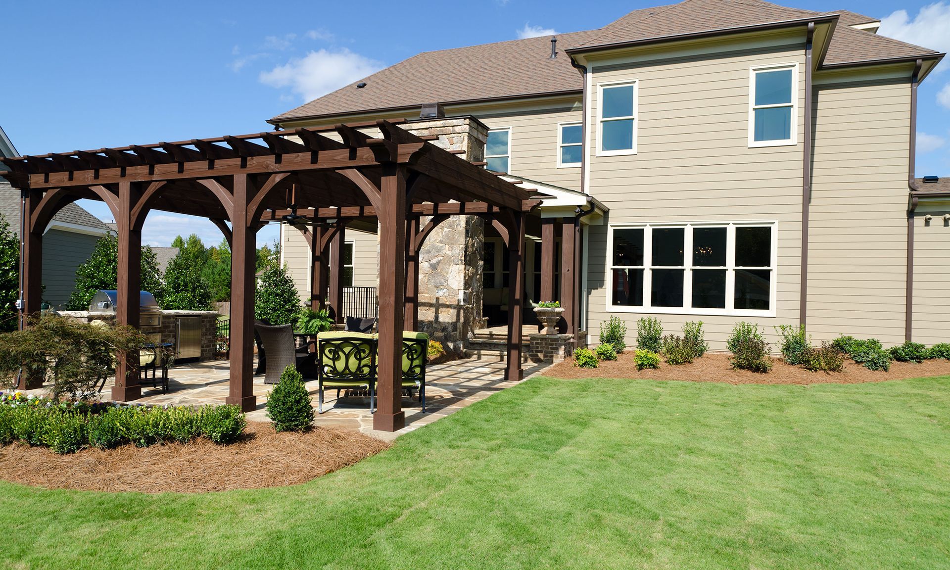 Backyard patio with wooden pergola beside a tan two-story house and green lawn.