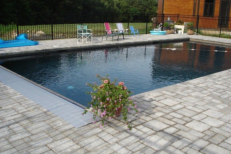 A rectangular swimming pool with dark water and a grey automatic cover, surrounded by stone pavers and lawn chairs.
