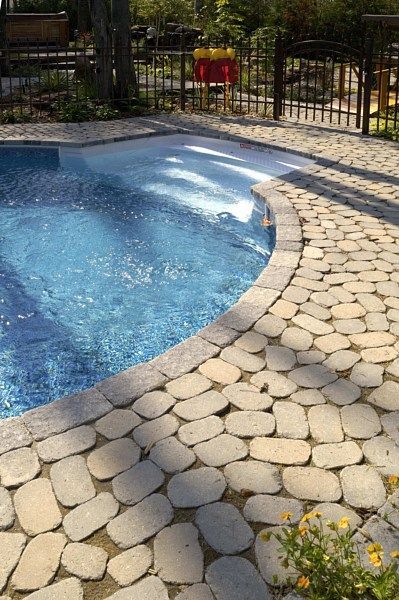 A curved swimming pool with light blue water and white steps, edged by a textured stone patio under bright sunlight.