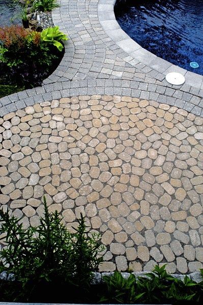 A circular patio made of rounded tan cobblestones, framed by a grey brick border next to a swimming pool and greenery.