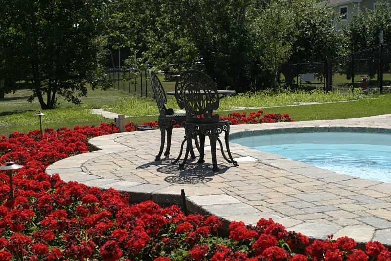 A metal table and two chairs sit on a paver patio next to a swimming pool, surrounded by vibrant red flowers.