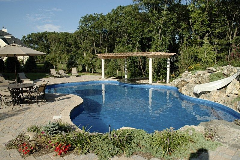 A curved swimming pool with a stone waterfall, a slide, and a white pergola on a patio surrounded by trees and landscaping.