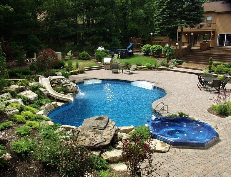 A backyard pool with a slide, stone rock features, a hot tub, and patio seating, set against a wooded landscape.