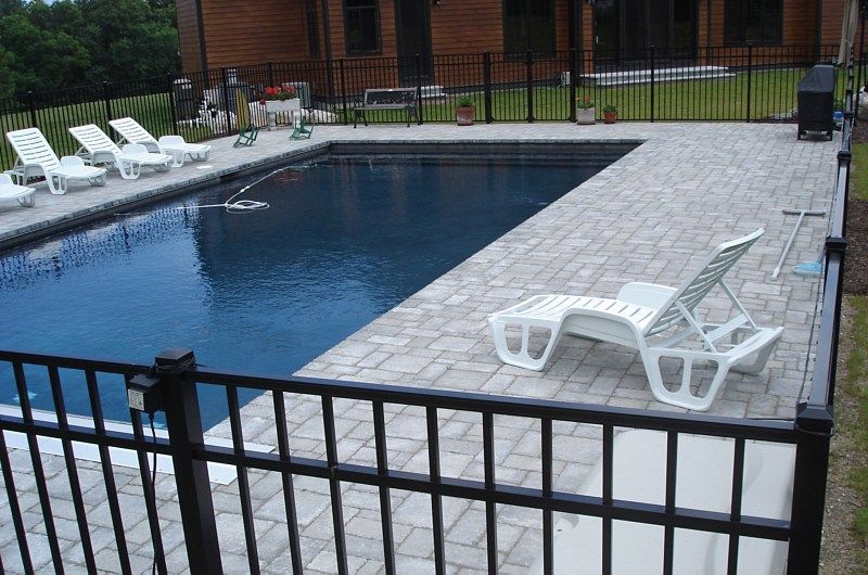 A rectangular swimming pool with blue water, surrounded by light-colored pavers, lounge chairs, and a black metal fence.