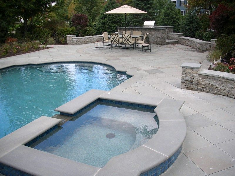 An outdoor patio with a swimming pool and hot tub, stone pavers, dining area, and umbrella, set in a backyard landscape.