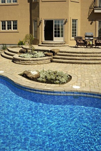 A beige house exterior with a patio, tiered stone steps, landscaped garden beds, and a bright blue swimming pool.