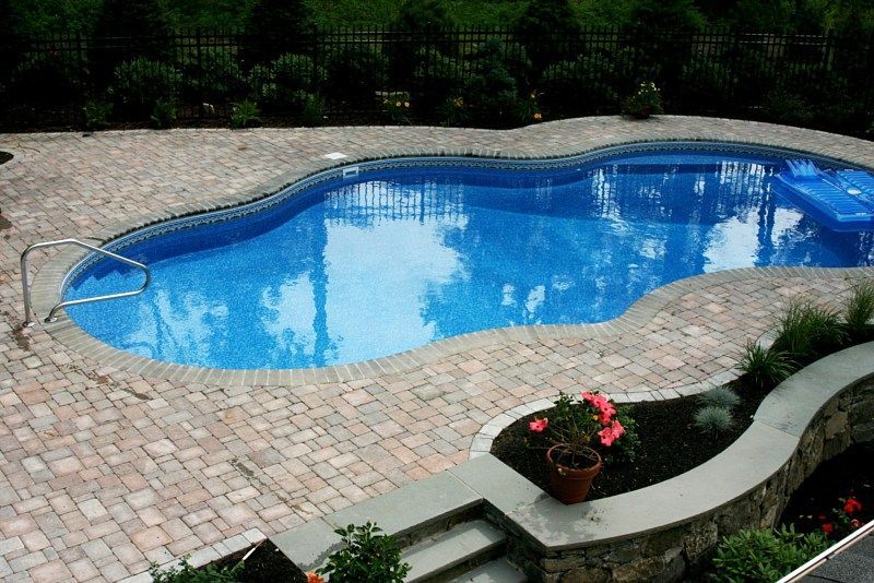 An irregularly shaped swimming pool with a blue tiled liner, surrounded by a light-colored paver patio and stone wall.