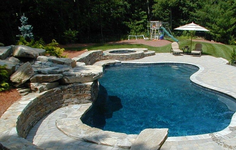 A kidney-shaped swimming pool with a stone rock feature, spa, and patio area in a backyard setting.