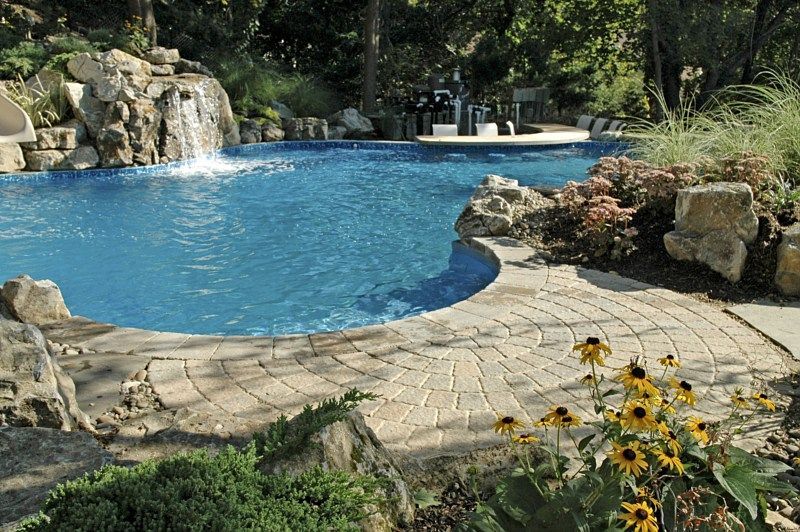 A backyard pool with a rock waterfall, blue water, and a paved circular patio surrounded by landscaping and flowers.