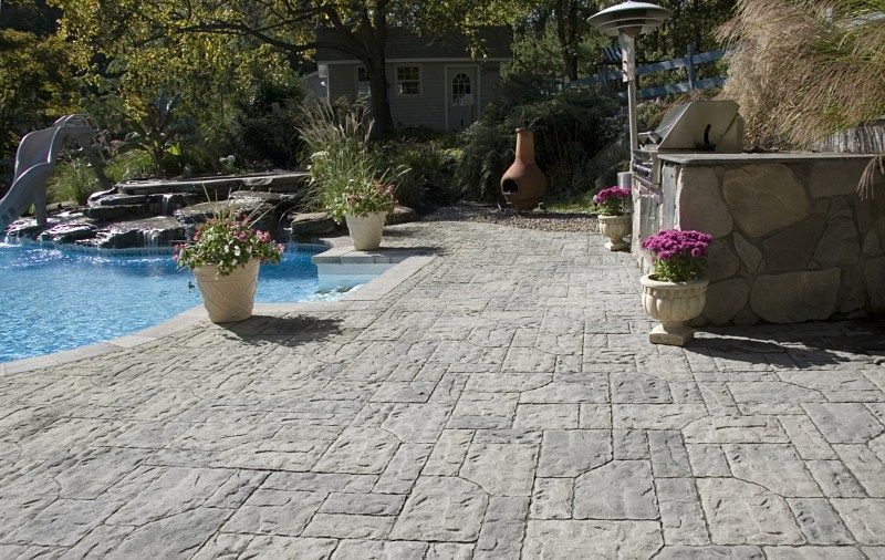 A stone patio overlooks a swimming pool with a slide, potted flowers, and an outdoor kitchen area.