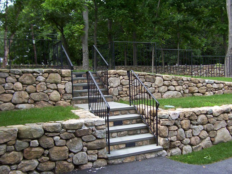 Stone retaining walls with built-in stairs featuring black iron railings, surrounded by grass and trees.