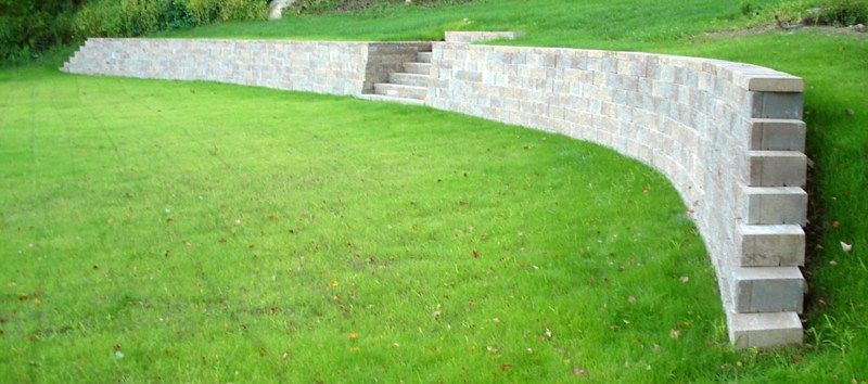 A curved stone retaining wall separates a grassy lawn from an upper grassy terrace, featuring built-in stone steps.