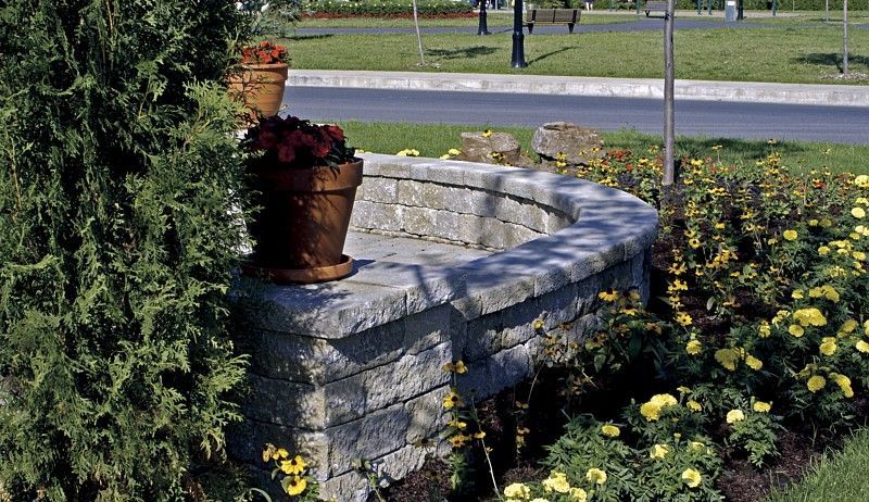 A stone retaining wall with potted plants and surrounding yellow flowers in a landscaped garden area.