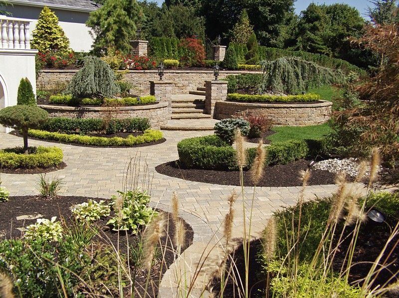 A sunny garden patio with tiered stone retaining walls, lush green shrubs, manicured trees, and decorative paved paths.