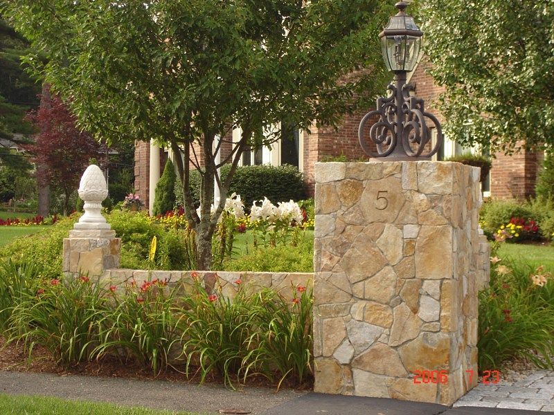 A stone pillar with the number 5, topped with an ornate lamp, next to a low stone wall and a white finial in a garden.