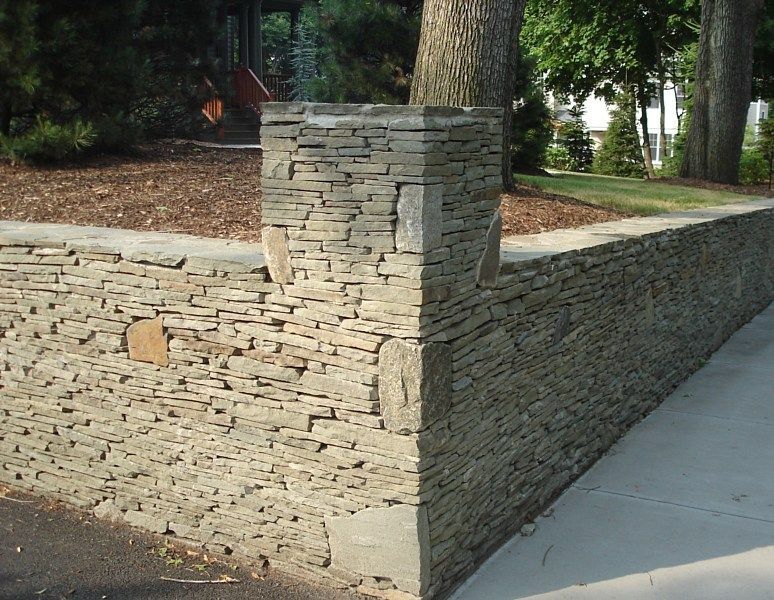 A corner section of a gray stacked stone retaining wall featuring a vertical pillar and a concrete walkway.
