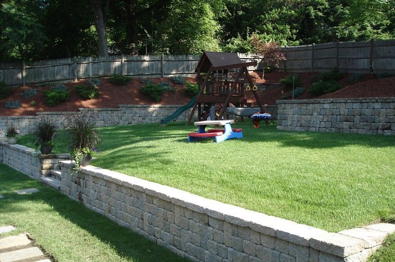A tiered backyard with a stone retaining wall, a grassy lawn, a small children’s play set, and a plastic picnic table.