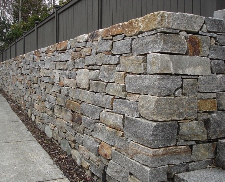 A sturdy, textured stone retaining wall constructed from stacked, multicolored gray and brown blocks next to a sidewalk.