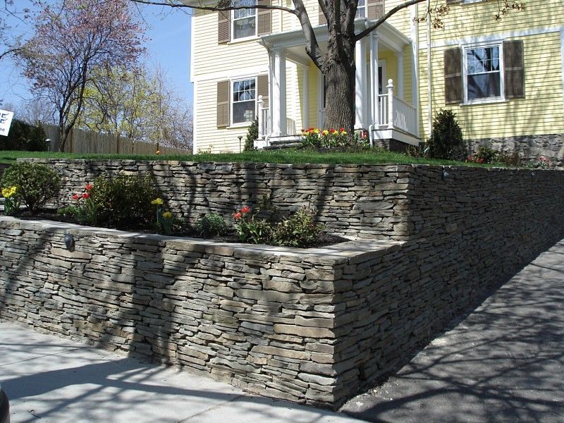 A tiered stone retaining wall with garden beds in front of a yellow house with a porch and mature tree.