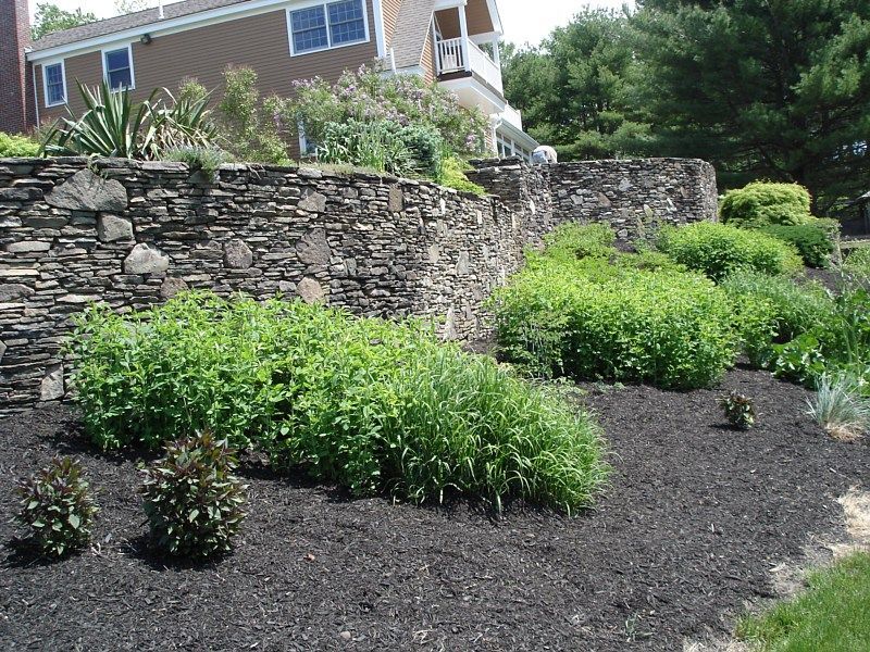 A stone retaining wall curves along a landscaped yard with lush green shrubs and dark mulch in front of a suburban home.