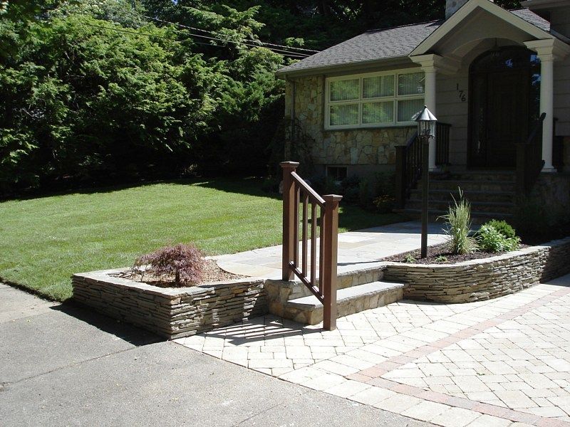 A stone-walled garden bed and steps leading to a house entrance, with a paved walkway and a small tree in the corner.