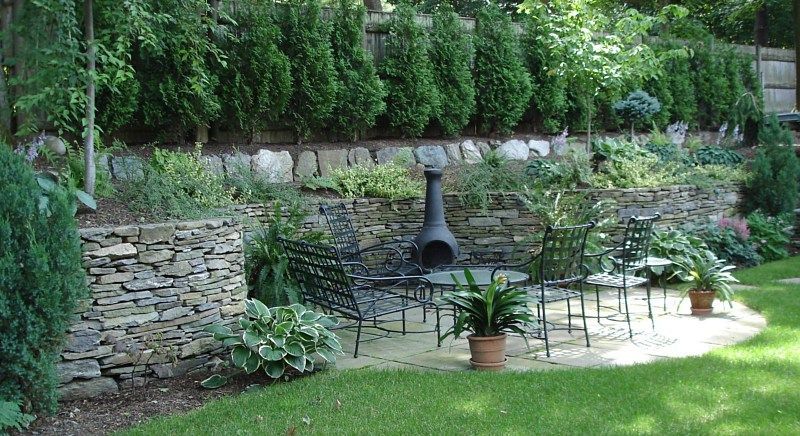 A stone patio area with a fire pit, metal chairs, and potted plants, set against a terraced wall and garden foliage.