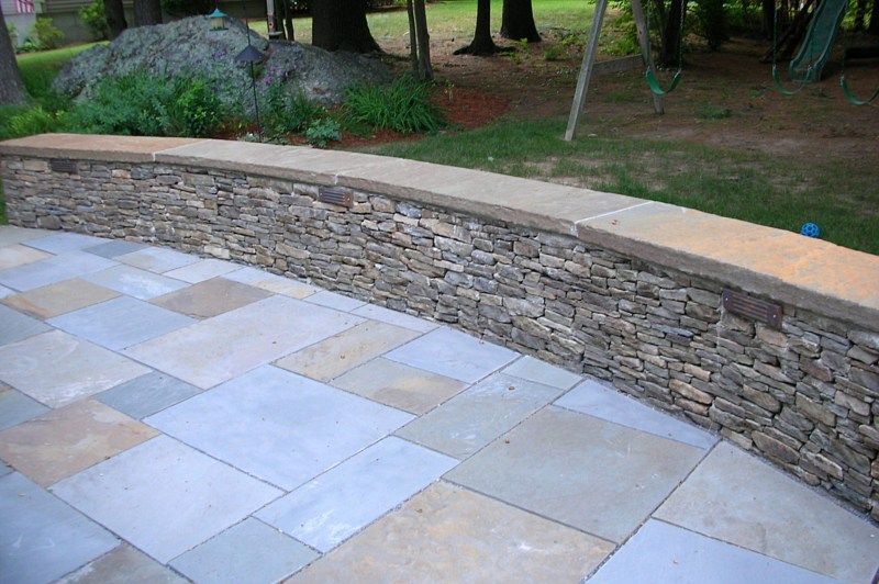 A curved stone retaining wall borders a multi-colored flagstone patio in a backyard setting.