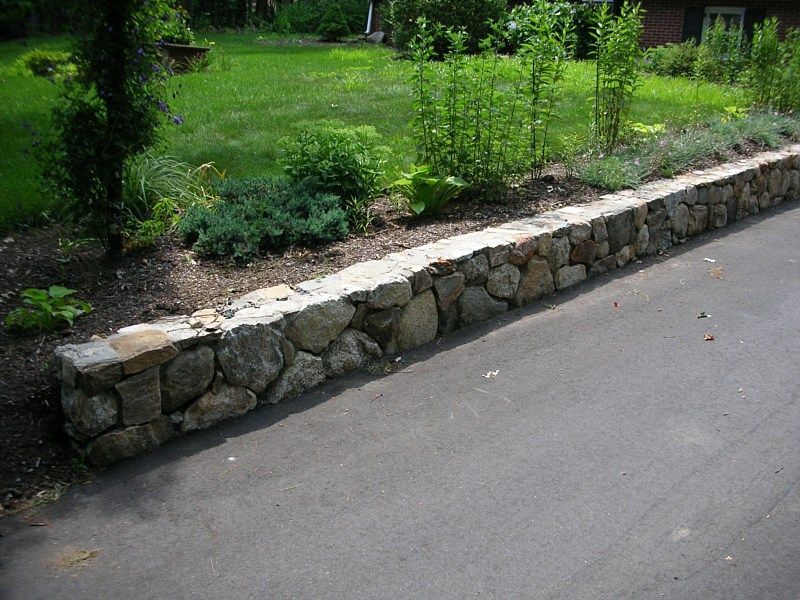 A low fieldstone retaining wall borders a paved driveway next to a grassy lawn with various shrubs and plants.