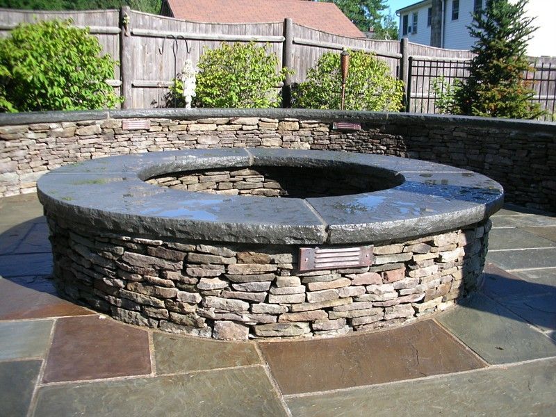 A circular stone fire pit with a dark, smooth stone top situated on a matching flagstone patio in a backyard.