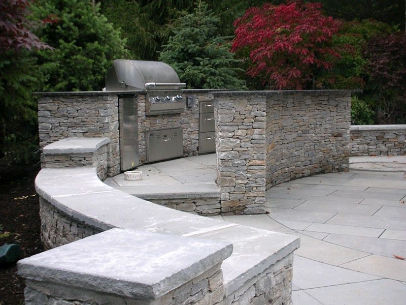 Outdoor stone kitchen island with a built-in grill and curved seating wall on a flagstone patio, surrounded by trees.