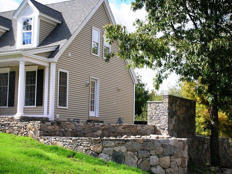 A beige house with white trim and dormer windows, featuring a natural stone patio and retaining wall in the front yard.