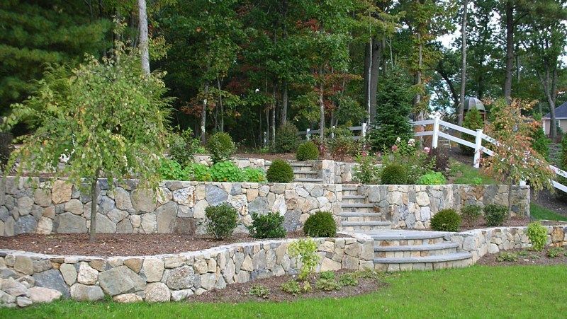 Tiered stone retaining walls with built-in stairs, landscaping, and a white fence in a garden setting.